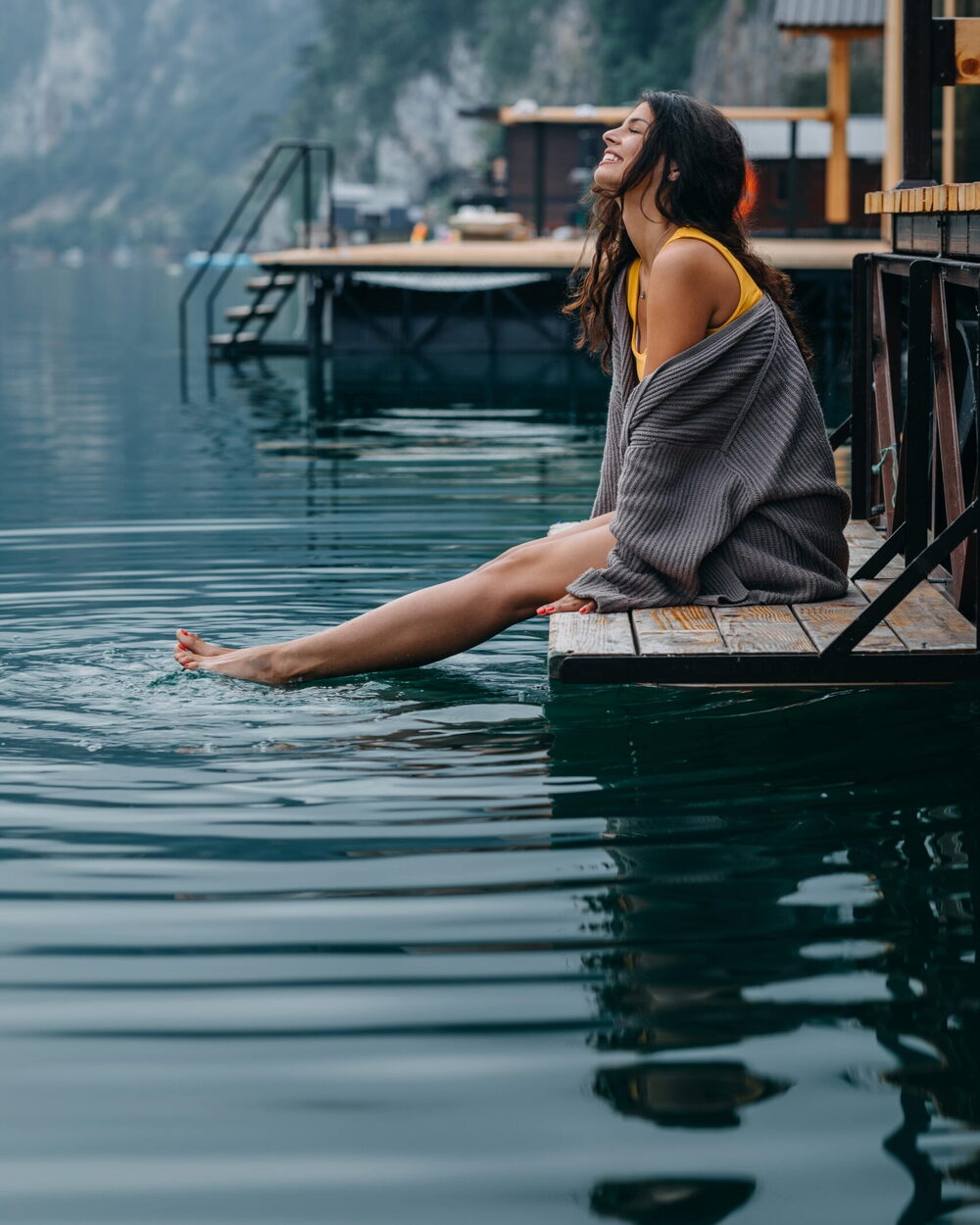Assise sur un ponton, une femme sourit les yeux fermés, les pieds dans l’eau d’un lac